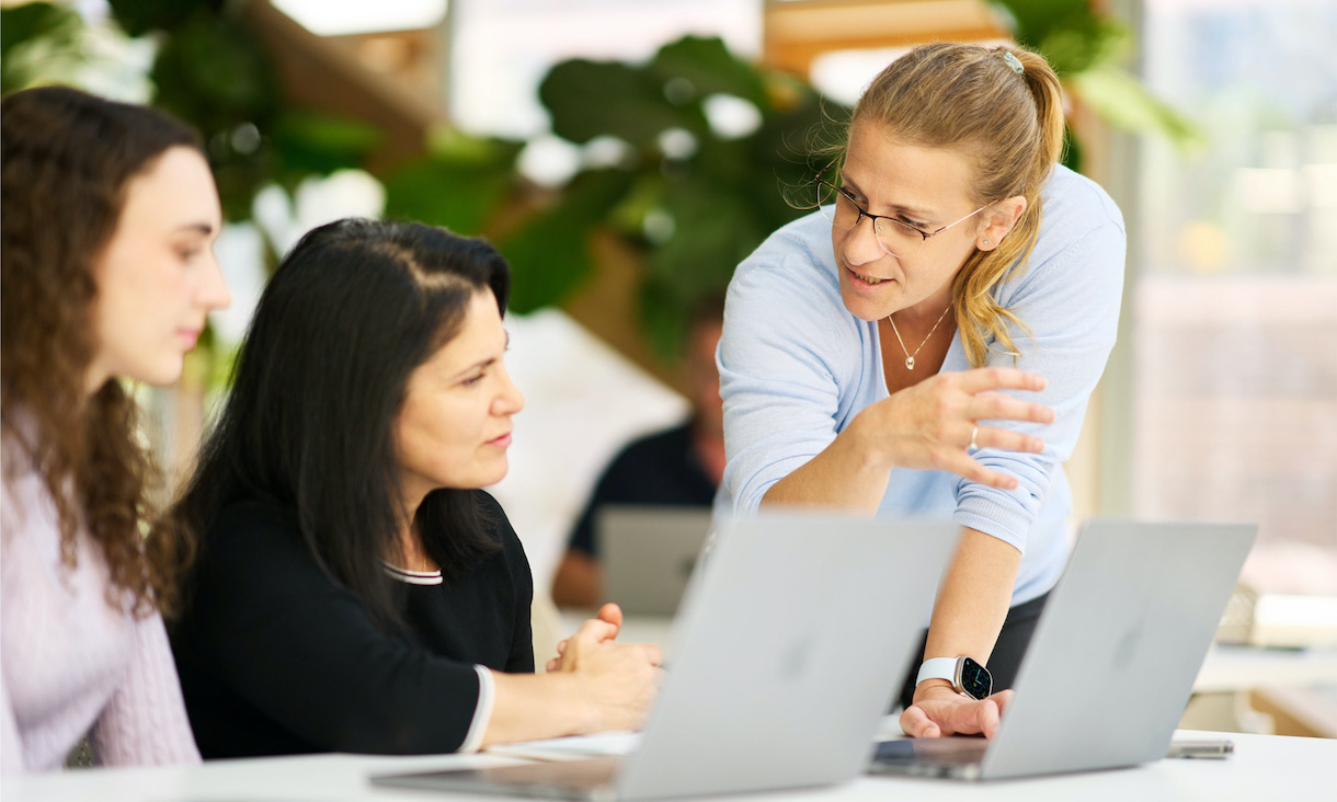 three women at laptops discussing their work, one is mentoring the other two