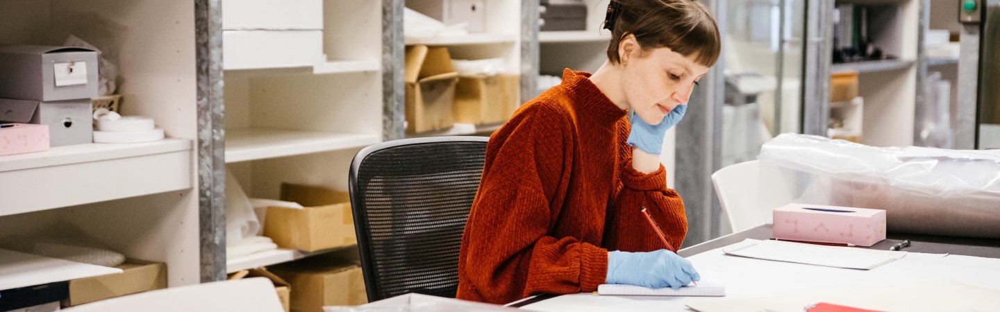 A photo of a woman sitting at a large table and writing on a notepad in the Design Archives building