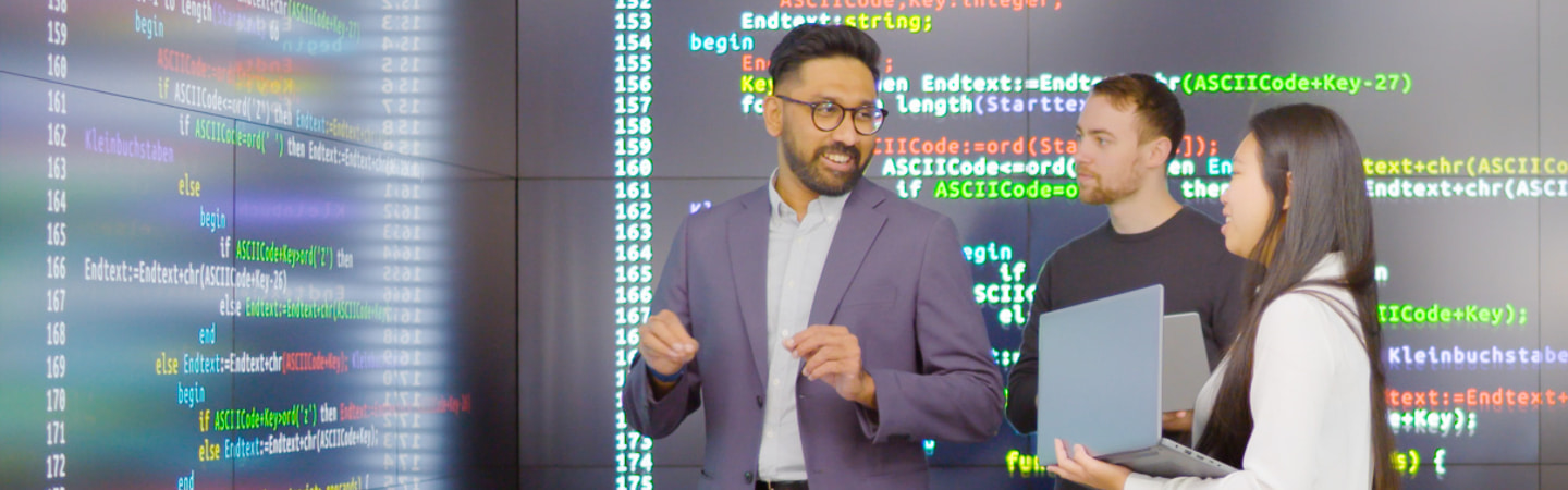 Two students holding laptops standing, looking towards a teacher, all standing in front of very large screens with lines of coloured computer code