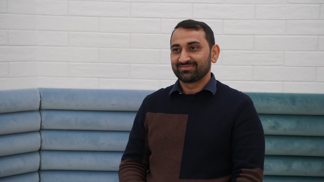 Nasir Mahmood wearing a dark jacket and sitting on a blue couch with a white brick wall behind them