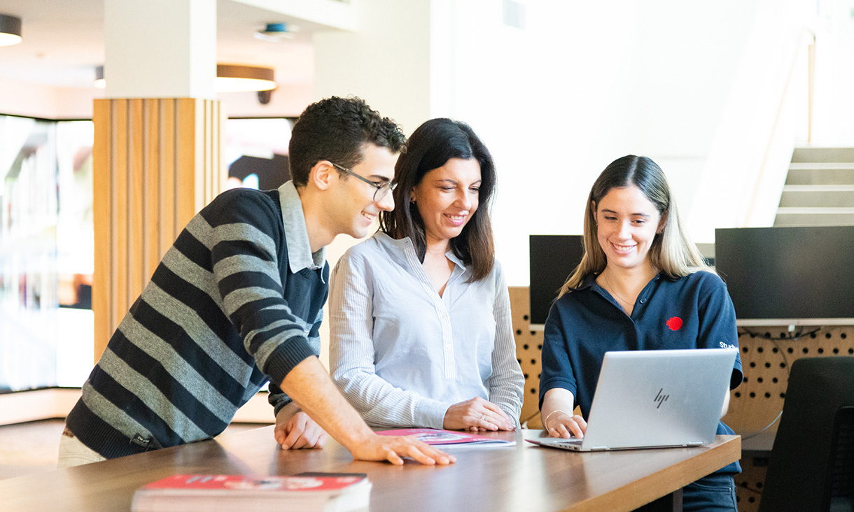 A parent with their teenaged child with a person wearing an RMIT shirt. The person in the RMIT shirt is showing something on their laptop screen.