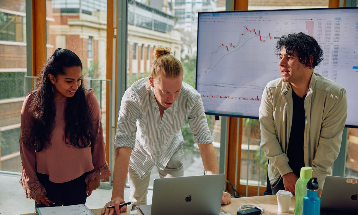 Three postgraduate students standing in front of a laptop on a desk, with a large screen behind them showing graph data, overlooking the RMIT campus on Bowen Street