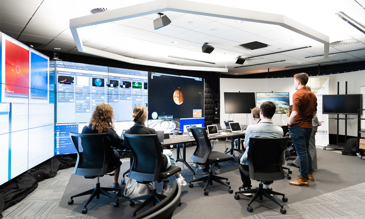 Group of students sitting and standing behind desks with laptops, looking towards very large screens