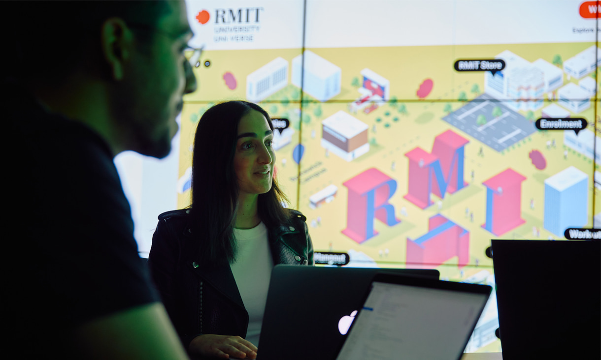 A female student in focus, and a male student in the foreground, both working on laptops, sitting in front of a large illuminated screen