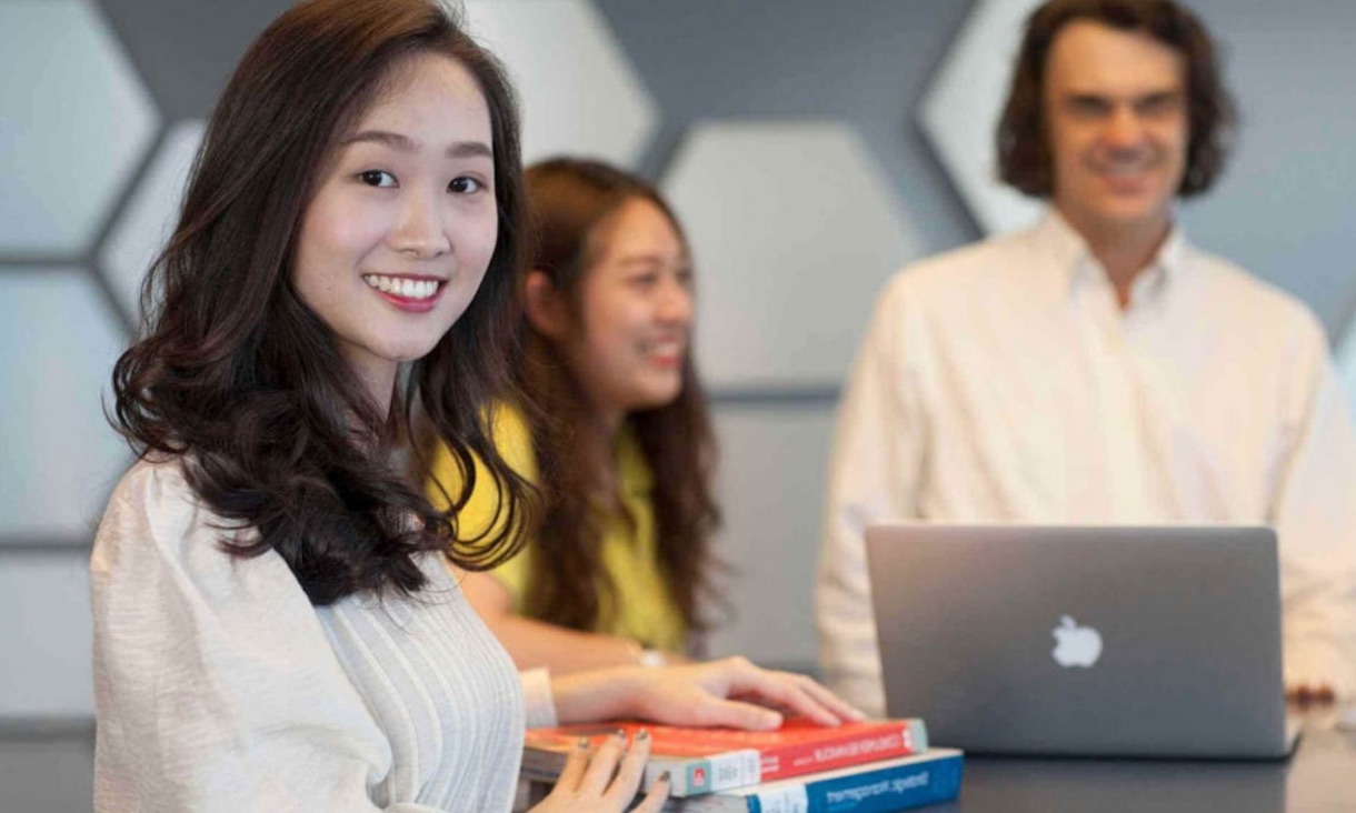 A student smiles at the camera while people work on a laptop behind her.